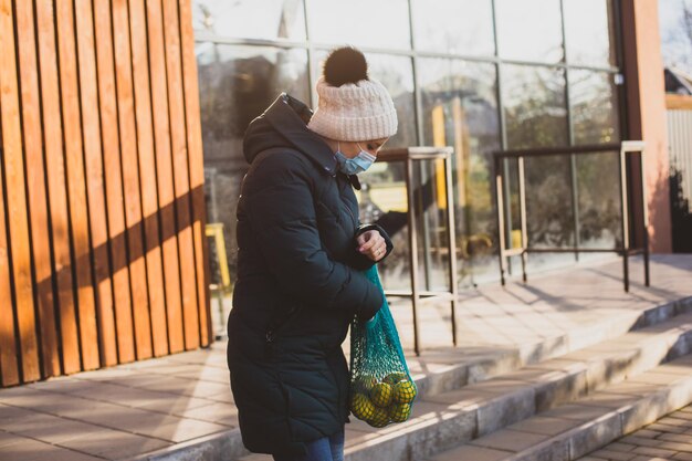 Donna in cappotto caldo e cappello lavorato a maglia dopo lo shopping tenendo una borsa a rete fatta a mano con frutta Soleggiata mattina d'inverno davanti al supermercato