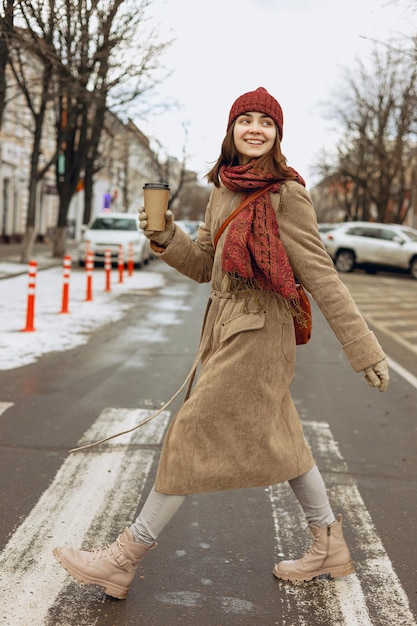 donna in cappotto e cappello caldo con sciarpa e tazza vuota di caffè da asporto attraversando la strada
