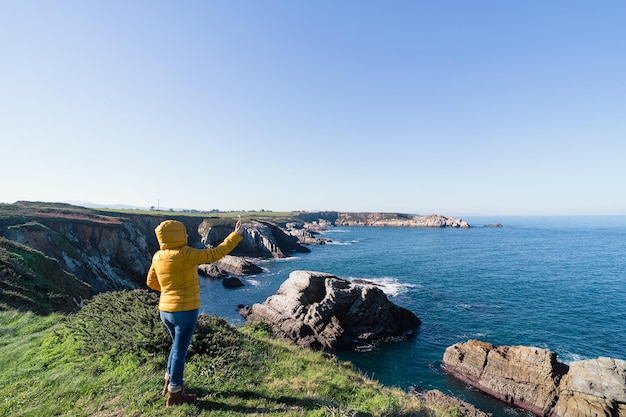 Donna in cappotto giallo che si fa un selfie in un paesaggio costiero naturale con mare e cielo blu Spazio di copia Foto orizzontale