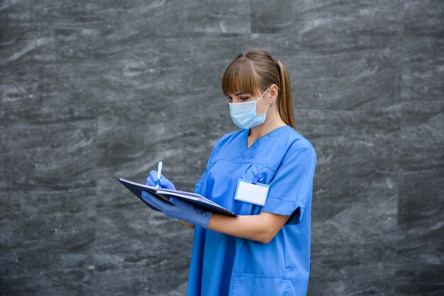 Donna in cappotto medico blu in posa su sfondo grigio in maschera con lo stetoscopio