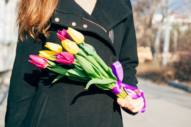 Donna in cappotto nero che tiene un mazzo di fiori luminosi in una giornata di sole