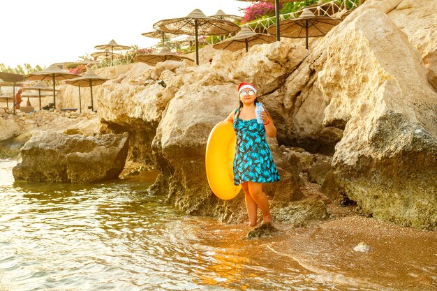 Donna in costume da bagno e cappello di Babbo Natale su una spiaggia rocciosa con un cerchio di nuoto e un cocktail.