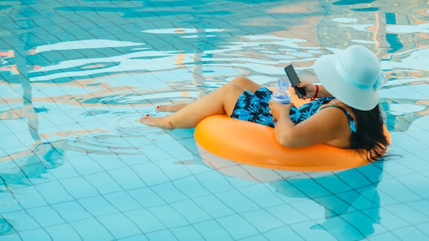 Donna in costume da bagno e cappello per il sole in piscina in un cerchio di nuoto con uno smartphone.