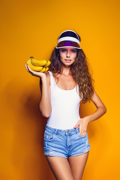 Donna in costume da bagno e shorts blu che tengono banana e posa isolati sopra la scena gialla