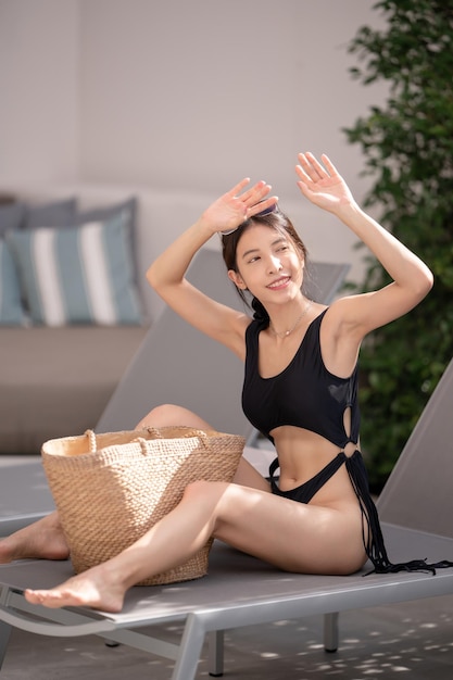 Donna in costume da bagno nero che tiene borsa da spiaggia in vimini di paglia fatta a mano vicino alla piscina