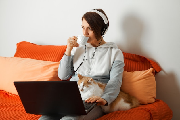 donna in cuffia con un computer portatile si siede sul letto e beve il caffè. Nelle vicinanze c'è un gatto che dorme sulla tastiera