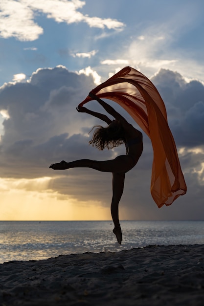 Donna in forma flessibile che salta con la seta durante il tramonto drammatico con nuvole tempestose concetto di aspirazione e opportunità