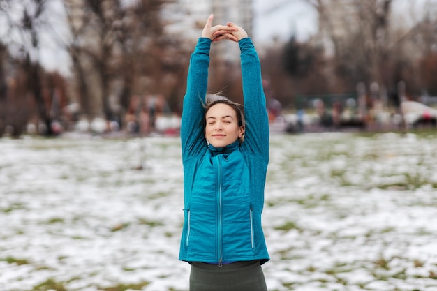 Donna in forma sportiva che fa esercizi di stretching per le braccia mentre si trova in un parco pubblico in una giornata invernale innevata. Fitness invernale, neve, clima freddo