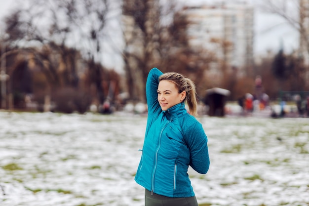 Donna in forma sportiva che fa esercizi di stretching per le braccia mentre si trova in un parco pubblico in una giornata invernale innevata. Fitness invernale, neve, clima freddo