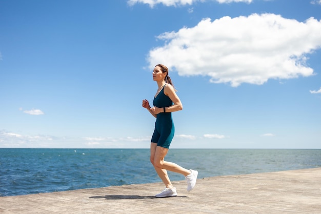 Donna in forma sportiva in abiti sportivi che fa jogging sul terrapieno in una luminosa giornata di sole con cielo blu e nuvole