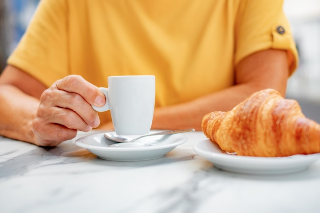 Donna in giallo con in mano una tazza di caffè espresso mentre è seduta al tavolo della mensa con un croissant fresco pronto da mangiare