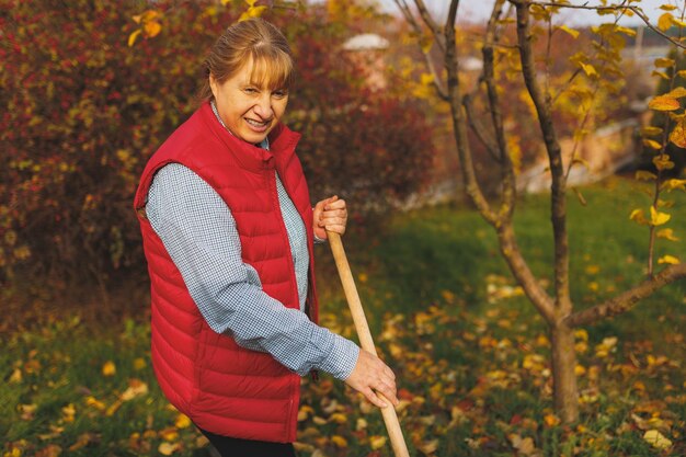 Donna in giubbotto rosso azienda rastrello Giardinaggio durante la stagione autunnale Pulizia del prato dalle foglie Rastrellare le foglie cadute nel giardino