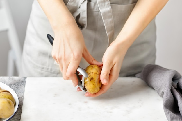 Donna in grembiule sbucciare patate bordo di marmo Mani sbucciare patate con pelapatate da cucina Preparazione del cibo