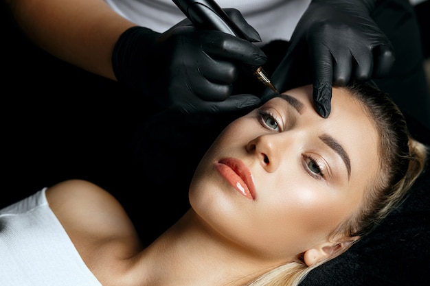 Donna in guanti che fa il trucco permanente della fronte a una bella donna bionda nel salone di cosmetologia. Vista dall'alto
