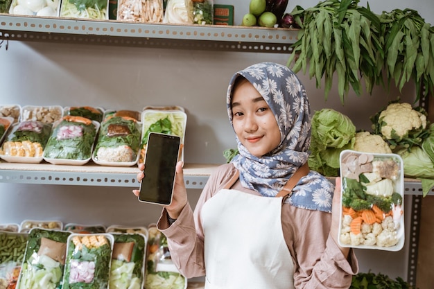 Donna in hijab che vende verdure che mostra le verdure e lo schermo mobile