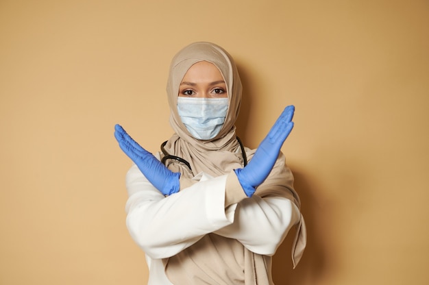 Donna in hijab e mascherina medica che gesturing esprimendo diniego e rifiuto sulla superficie beige con lo spazio della copia