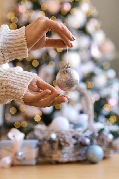 Donna in maglione di lana caldo bianco che tiene la palla decorativa del giocattolo in mani sui precedenti di un albero di Natale. Natale, concetto di celebrazione delle vacanze di capodanno. Messa a fuoco selettiva