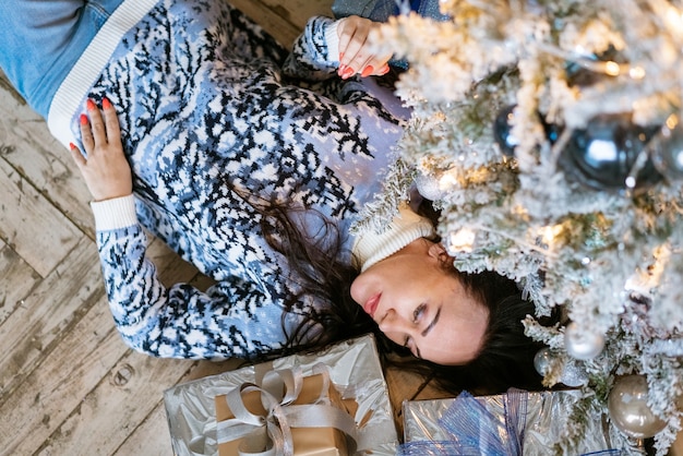 Donna in maglione di natale lavorato a maglia sdraiato sul pavimento vicino all'albero di natale godendo il festoso giorno invernale brun...