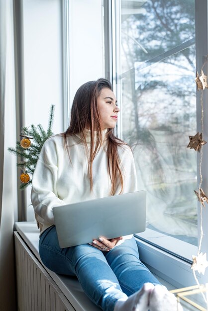 Donna in maglione invernale bianco caldo seduto al davanzale di casa alla vigilia di natale che lavora al computer portatile