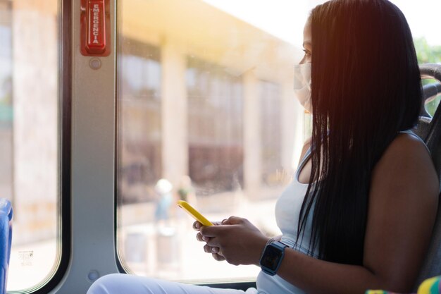 Donna in maschera protettiva che consulta il cellulare e guarda fuori dalla finestra mentre guida un autobus in città durante un'epidemia