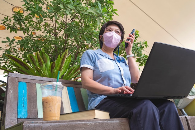 Donna in maschera protettiva che parla su smartphone e usa il laptop su una panca di legno nel centro commerciale