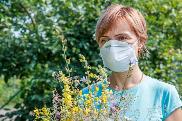 Donna in maschera protettiva che tiene un mazzo di fiori di campo e cerca di combattere le allergie al polline. Donna che protegge il naso dagli allergeni. Concetto di allergia.