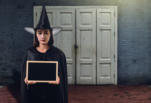 Donna in nero spaventoso strega costume di halloween, tenendo lavagna con muro di mattoni di Ancie
