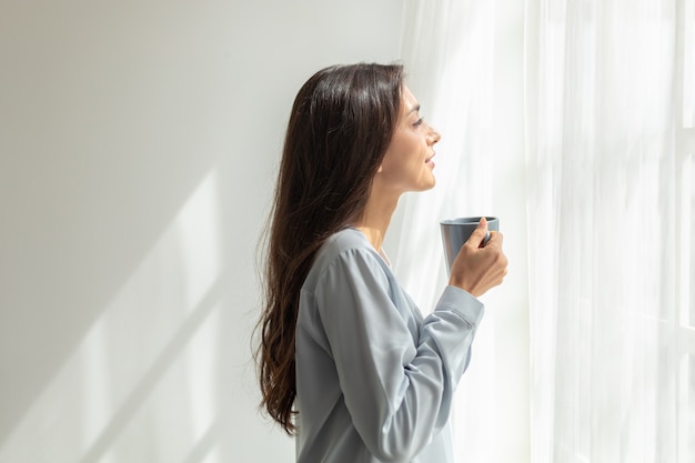 donna in piedi beve caffè e apre le tende della finestra respira aria fresca esercizio di stiramento in camera da letto