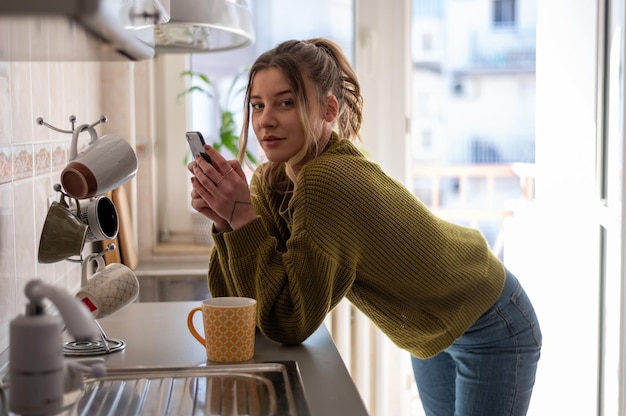 Donna in piedi in cucina e bere una tazza di tè mentre utilizza lo smartphone