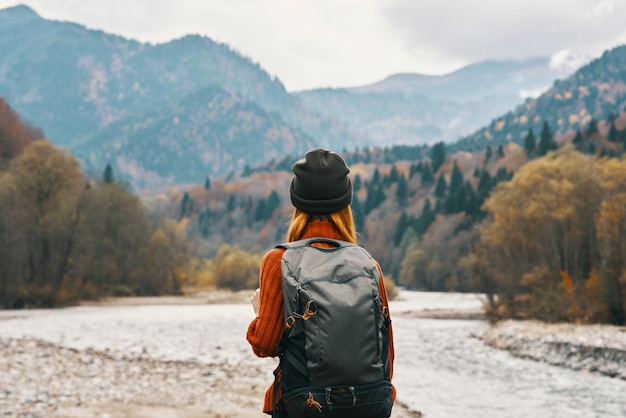 Donna in piedi sulla riva del fiume e guarda le montagne in lontananza paesaggio autunno zaino modello di turismo foto di alta qualità