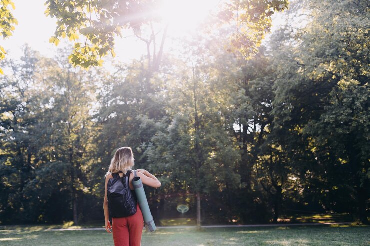 Foto donna in piedi vicino agli alberi in una giornata di sole che porta un tappetino da yoga
