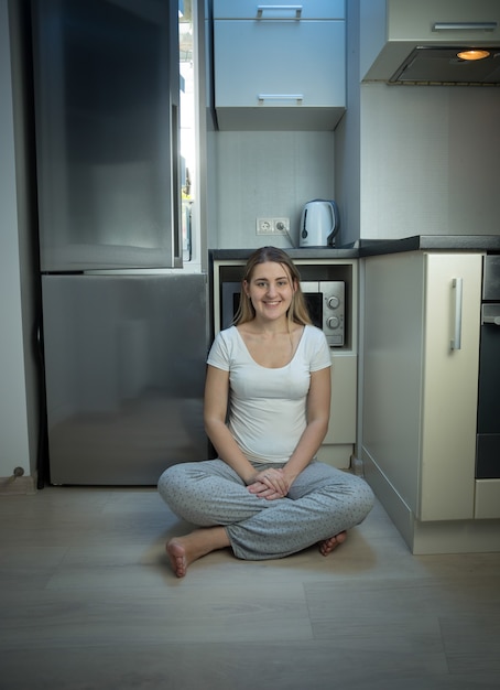 Donna in pigiama seduta sul pavimento in cucina accanto al frigorifero aperto a tarda sera