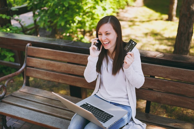 Donna in possesso di carta di credito, parlando al telefono cellulare, avendo una conversazione di buon piacere. Donna seduta su una panchina che lavora al computer portatile moderno in strada all'aperto. Ufficio Mobile. Concetto di business freelance.