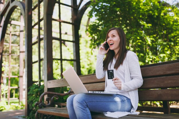 Donna in possesso di carta di credito, parlando al telefono cellulare, avendo una conversazione di buon piacere. Donna seduta su una panchina che lavora al computer portatile moderno in strada all'aperto. Ufficio Mobile. Concetto di business freelance.