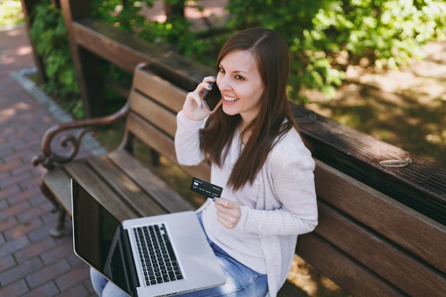 Donna in possesso di carta di credito, parlando al telefono cellulare, avendo una conversazione di buon piacere. Donna seduta su una panchina che lavora al computer portatile moderno in strada all'aperto. Ufficio Mobile. Concetto di business freelance.