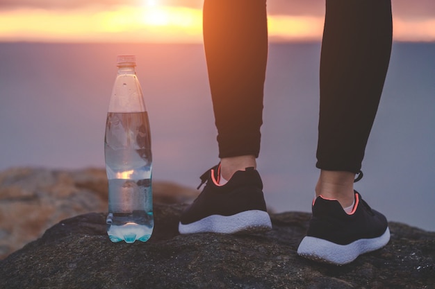 Donna in scarpe da ginnastica in piedi su una roccia accanto a una bottiglia di acqua su uno sfondo di mare al tramonto