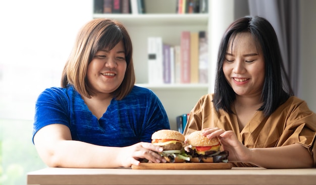 Donna in sovrappeso affamata che tiene hamburger su un piatto di legno, durante il lavoro da casa, aumenta il problema di peso. Concetto di disturbo da alimentazione incontrollata (BED).