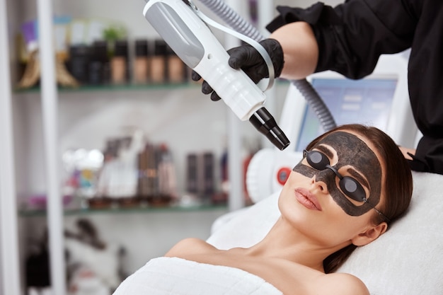 Donna in spa con maschera di carbonio nero e cosmetologo con laser vicino al viso, copia spazio, bella donna con ringiovanimento da estetista