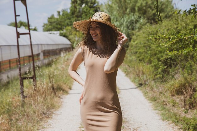 donna in un abito beige e cappello di paglia in posa su una strada di campagna