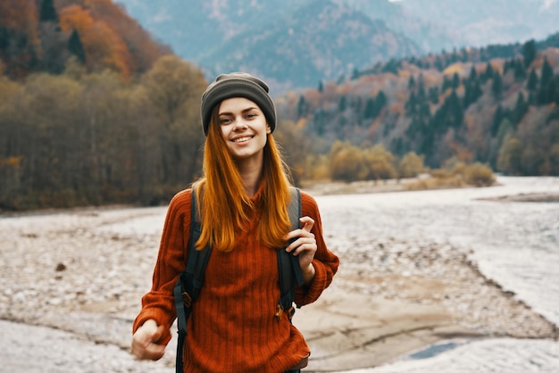 Donna in un cappello da maglione con uno zaino che riposa vicino al fiume in montagna sul paesaggio naturale
