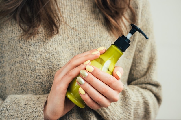 Donna in un maglione marrone con il manicure giallo che tiene una bottiglia verde con crema cosmetica, primo piano