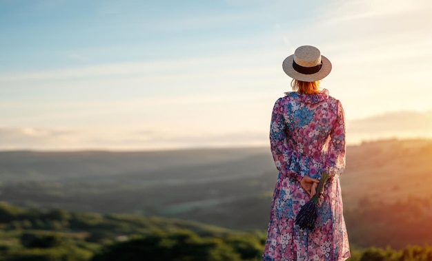 Donna in un vestito e un cappello con un bouquet di lavanda nelle sue mani su una collina nel tramonto estivo