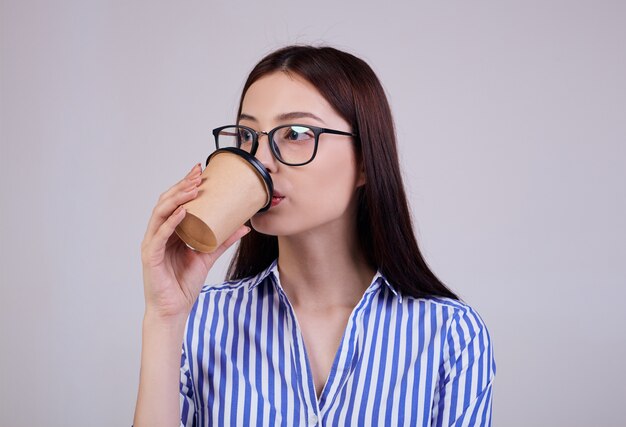 donna in una camicia a righe e occhiali pc nero sta con una tazza di caffè marrone in mano su grigio. La donna guarda con calma dritto. Bevande.