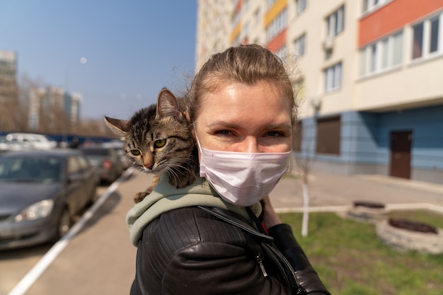 donna in una maschera medica sta camminando con un gatto lungo la strada vicino alla casa