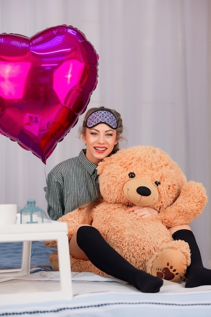 donna in una maschera per dormire sorride e abbraccia un orso giocattolo il giorno di San Valentino, accanto a un cuore di palloncino rosa