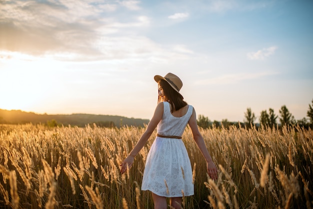 Donna in vestito bianco e cappello di paglia che cammina nel campo di grano sul tramonto. Persona di sesso femminile sul prato estivo, vista posteriore