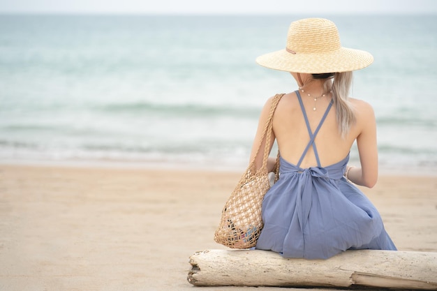 Donna in vestito blu e cappello di paglia che si siede su un legname dall'oceano