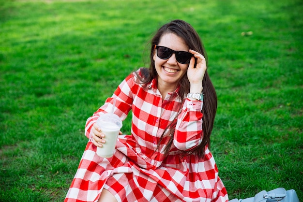 Donna in vestito rosso che beve frullato mentre è seduto sull'erba verde nel parco