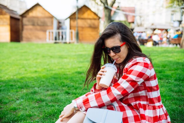 Donna in vestito rosso che beve frullato mentre è seduto sull'erba verde nel parco