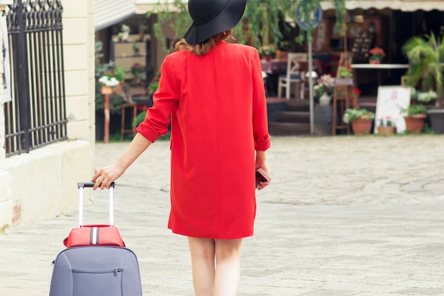 Donna in viaggio con cappello nero che cammina con la valigia in strada della città.